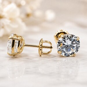 May include: A pair of stud earrings featuring round, clear, faceted stones set in yellow gold prongs. One earring is shown from the side, revealing the post and threaded screw back, while the other is positioned to display the circular stone face.