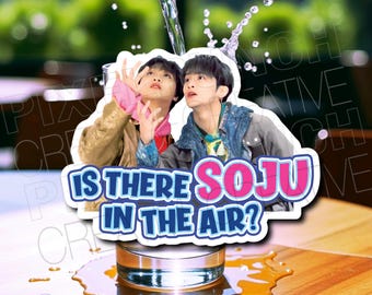 May include: A sticker with two men in a humorous pose, with the text "Is there Soju in the air?" in pink and blue lettering. The sticker is placed on a glass of water with a brown wooden surface in the background.
