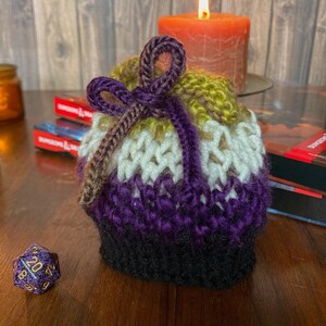 May include: A crocheted drawstring bag with a purple, white, yellow, and brown color scheme. The bag is tied with a purple ribbon and is sitting on a wooden surface.