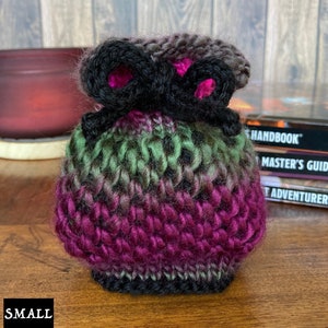 May include: A small, crocheted drawstring bag with a black and pink color scheme. The bag has a black ribbon tied around the top and is sitting on a wooden surface.