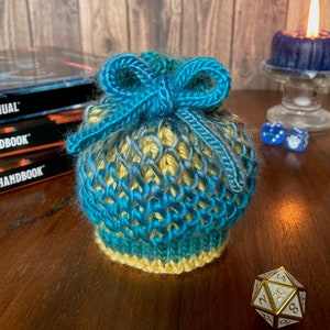 May include: A blue and yellow knitted dice bag with a blue ribbon tied in a bow. The bag is sitting on a wooden table with a gold metal dice and two blue dice.