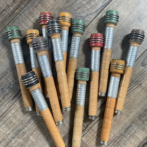 May include: A collection of vintage wooden spools with metal and colored accents. The spools have a natural wood finish with silver metal bands and tops in various colors, including red, green, and black. The spools are arranged on a wooden surface.