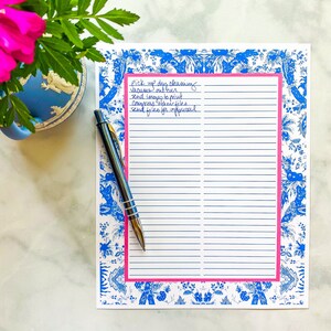 May include: A white notepad with blue and white floral border and pink accents. The notepad has lined pages and a handwritten to-do list that reads: "Pick up dry cleaning, Vacuums out boxes, Send images to print, Congress office files, Send files for approval."