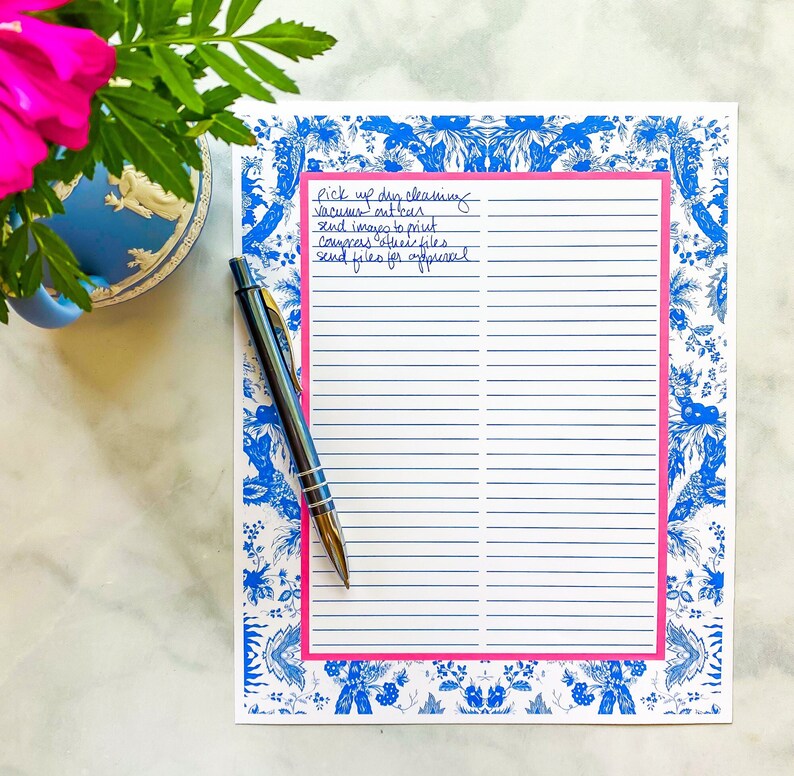 May include: A white notepad with blue and white floral border and pink accents. The notepad has lined pages and a handwritten to-do list that reads: "Pick up dry cleaning, Vacuums out boxes, Send images to print, Congress office files, Send files for approval."