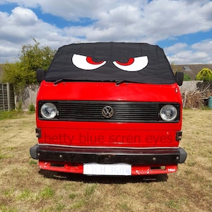 Może przedstawiać: Czerwony samochód dostawczy Volkswagen z czarną osłoną przeciwsłoneczną na przednią szybę, na której znajdują się dwa duże białe oczy z czerwonymi źrenicami. Samochód ma czarny przedni zderzak i czarną kratkę z logo Volkswagena. Na przednim zderzaku widoczny jest napis "betty blue screen eyes".