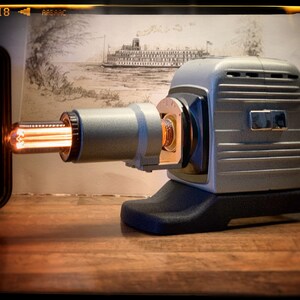 Könnte beinhalten: Ein vintage silberner Diaprojektor mit schwarzem Sockel und einer leuchtend orangefarbenen Glühbirne. Der Projektor steht auf einer Holzoberfläche.