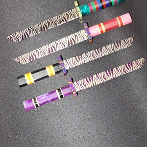 May include: Four miniature swords with zebra-striped blades and colorful handles. The handles are made of different materials, including wood, metal, and plastic. The swords are arranged in a row on a dark gray surface.