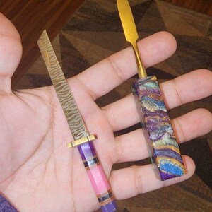 May include: Two small knives with colorful handles. The first knife has a gold blade and a purple, blue, and gold handle. The second knife has a silver blade with a pink, purple, and gold handle.