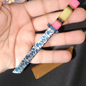 May include: A small, blue and silver pocket knife with a pink, yellow, and black handle. The knife has a decorative pattern on the blade.