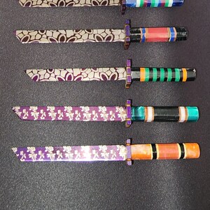 Könnte beinhalten: Sechs Miniaturschwerter mit violetten Griffen und dekorativen goldenen Designs auf den Klingen. Jedes Schwert hat einen andersfarbigen Griff mit schwarzen und goldenen Streifen.