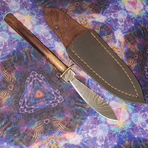 May include: A small, handcrafted knife with a wooden handle and a patterned steel blade. The knife is in a brown leather sheath.