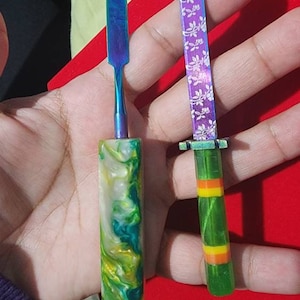 Könnte beinhalten: Zwei kleine Werkzeuge mit bunten Griffen. Das Werkzeug links hat einen grün-weiß-gelb marmorierten Griff und eine blaue Metallklinge. Das Werkzeug rechts hat einen grünen Griff mit orange-gelben Streifen und eine violette Metallklinge mit einem weißen Blumenmuster.