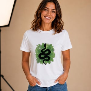 May include: A white short-sleeved t-shirt featuring a central graphic print of a black snake coiled amidst a cluster of vibrant green tropical leaves. The design is positioned on the chest area of the garment, which has a crew neckline.