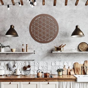 May include: A circular brown wall art piece featuring a geometric flower of life pattern with intersecting circles. The wall decor is mounted on a gray textured wall above a kitchen counter with various kitchenware items and hanging pendant lights.