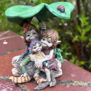 May include: Three small green and brown fairy figurines are huddled together under a green shamrock leaf. A red ladybug is perched on the leaf. The fairies are wearing colorful clothing and have large, expressive eyes.