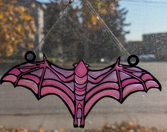 Puede incluir: Un murciélago de vidrio tintado de color rosa que cuelga de una ventana con una ventosa. El murciélago tiene un diseño detallado con un contorno negro.