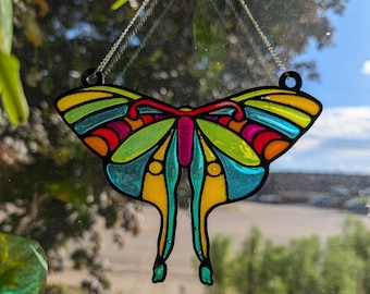 Puede incluir: Un adorno de mariposa de vidrio coloreado con detalles en amarillo, verde, azul, rojo y rosa. La mariposa está colgada de una cadena y suspendida frente a una ventana.