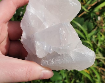 May include: A translucent, white crystalline mineral specimen with a jagged, multi-faceted surface. The cluster features sharp, angular edges and a semi-clear, glass-like texture that catches the sunlight, displaying internal fractures and natural geometric formations throughout the stone.