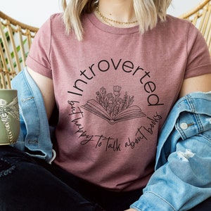 Puede incluir: Una camiseta color malva con un dise&ntilde;o gr&aacute;fico negro que dice "Introvertido pero feliz de hablar de libros". El gr&aacute;fico presenta un libro con flores que salen de &eacute;l.