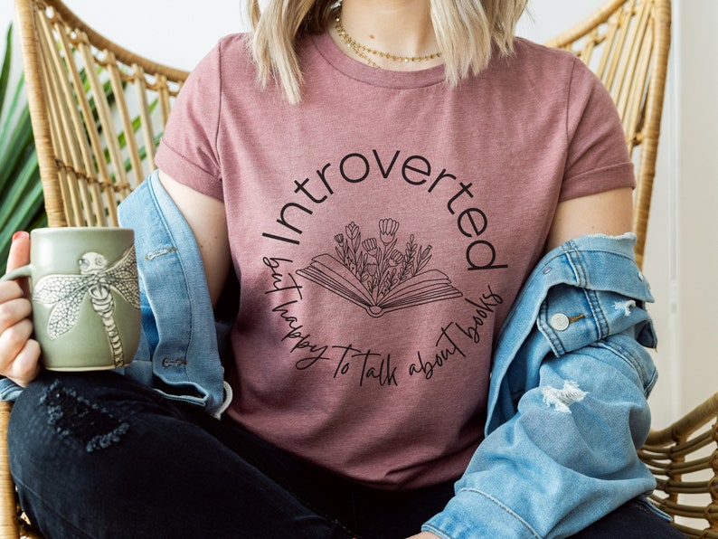 Puede incluir: Una camiseta color malva con un dise&ntilde;o gr&aacute;fico negro que dice "Introvertido pero feliz de hablar de libros". El gr&aacute;fico presenta un libro con flores que salen de &eacute;l.