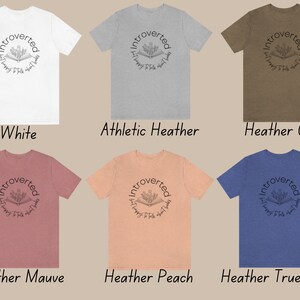 Puede incluir: Seis camisetas de diferentes colores con el texto "Introvertido y feliz de hablar de libros" en un c&iacute;rculo alrededor de un dibujo de un libro abierto con flores que salen de &eacute;l.