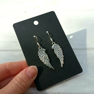 May include: A pair of silver dangle earrings with a wing design. The earrings are displayed on a black card.