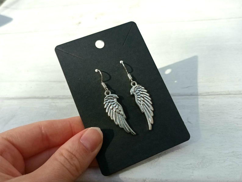 May include: A pair of silver dangle earrings with a wing design. The earrings are displayed on a black card.
