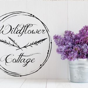 May include: A white background features a black circular design with the words "Wildflower Cottage" in elegant script. A bouquet of purple lilacs sits in a small, galvanized metal bucket, adding a touch of color and freshness to the scene.