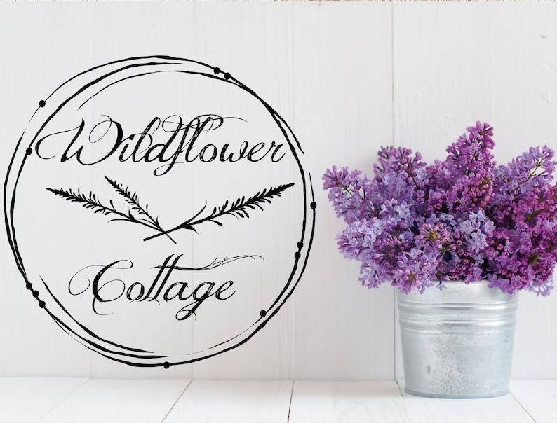 May include: A white background features a black circular design with the words "Wildflower Cottage" in elegant script. A bouquet of purple lilacs sits in a small, galvanized metal bucket, adding a touch of color and freshness to the scene.