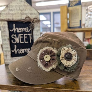 May include: A brown military-style cap with two fabric flower embellishments. One flower is brown with white polka dots and a beige button, the other is green with a cream-colored lace center and a dark brown button. The cap has distressed details.