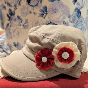 May include: A tan-colored cap with a short brim, adorned with two fabric flower embellishments. One flower is red, the other is cream, both with white button centers. The cap is set against a floral background.