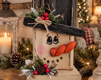 May include: A wooden snowman decoration featuring a black top hat, a painted face with pink cheeks, and a protruding orange carrot nose. The piece is adorned with artificial greenery, red berries, and a small twine bow.