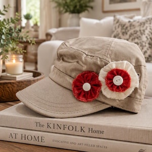 May include: A tan military-style cap featuring two fabric flower embellishments on the side. One flower is solid red with a white center button, and the other is white with a red center and a white button. The cap has a structured brim.