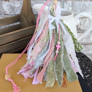 May include: A handmade tassel ornament with pink, green, and white fabric strips, lace, and a wooden bead. A pink cord and a small pink cross charm with pearls are attached. The tassel is resting on a book.