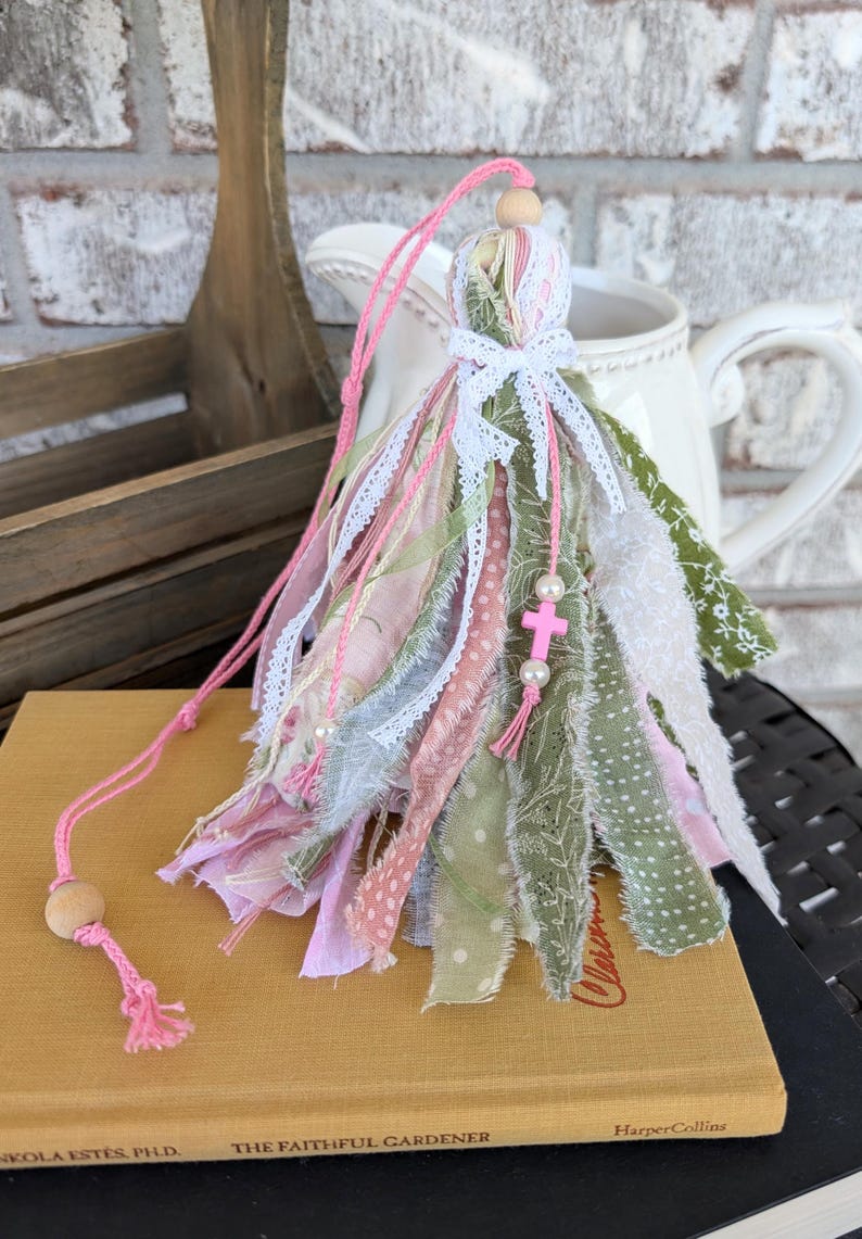 May include: A handmade tassel ornament with pink, green, and white fabric strips, lace, and a wooden bead. A pink cord and a small pink cross charm with pearls are attached. The tassel is resting on a book.
