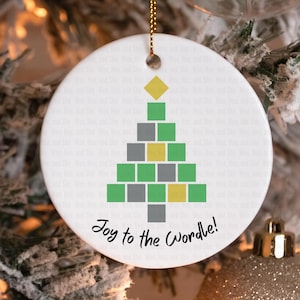 May include: A white ceramic Christmas ornament with a gold string for hanging. The ornament features a pixelated Christmas tree design in green, gray, and yellow squares, with a yellow square at the top. The text "Joy to the Wordle!" is written below the tree.