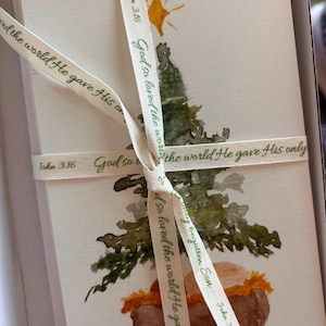 Puede incluir: Un montón de tarjetas navideñas de acuarela atadas con una cinta. Cada tarjeta presenta una pintura de acuarela de un árbol de Navidad y una escena del pesebre. La cinta tiene el texto "Porque de tal manera amó Dios al mundo, que ha dado a su Hijo unigénito" y "Juan 3:16".