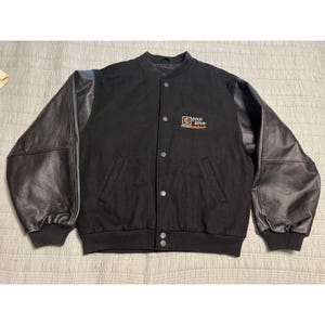 May include: Black varsity jacket with black leather sleeves. The jacket has a button-up front, two front pockets, and a ribbed collar and cuffs. The Ditch Witch logo is on the upper left chest.