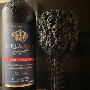 May include: A black wine glass with a skull design. The glass is filled with a dark liquid. The glass is sitting on a table next to a bottle of Stella Rosa Blueberry Reserve wine.