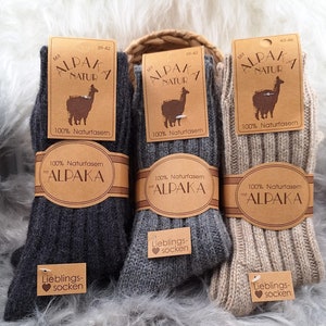 Peut inclure: Trois paires de chaussettes en laine d'alpaga. Les chaussettes sont en gris, beige et marron. Chaque paire a une étiquette qui dit "Alpaca Natur" et "Lieblings-Socken".