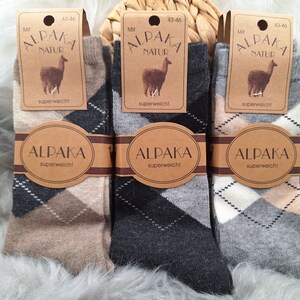 Op de afbeelding: Drie paar sokken met een ruitpatroon in beige, zwart en grijs. De sokken zijn gemaakt van alpaca wol en hebben een label met de tekst "Alpaca Natur" en "superweich!" wat "superzacht!" betekent in het Duits.