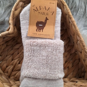 Könnte beinhalten: Ein Paar beigefarbene Alpaka-Socken mit einem Etikett, auf dem "Mit Alpaka Natura 43-46 100% superweich!" steht und ein Alpaka-Cartoon abgebildet ist.