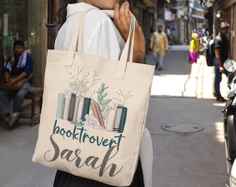 May include: A beige tote bag with a floral design and the text "booktrovert Sarah". The bag features a stack of colourful books with flowers and leaves around them.