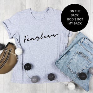 May include: A gray t-shirt with the word "Fearless" printed on the front. The back of the shirt says "On the back: God's got my back". The shirt is laid out on a white wooden surface with blue jeans, a brown bag, and white and black decorative balls.