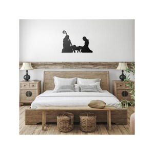 May include: A black metal silhouette of the nativity scene with Mary, Joseph, and the baby Jesus in a manger. The silhouette is mounted on a white wall above a wooden bed with a white duvet and pillows. Two wooden bedside tables with lamps are on either side of the bed.