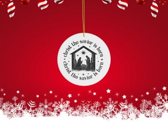 May include: A white, round Christmas ornament with a black and white nativity scene illustration. The text "Christ the savior is born" encircles the image. The ornament hangs from a gold string against a red background with Christmas decorations.