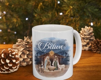 May include: White ceramic mug featuring a nativity scene with the word "Believe" in elegant script. The design includes a star, figures, and animals. The mug is set against a blurred background of pine needles and lights.