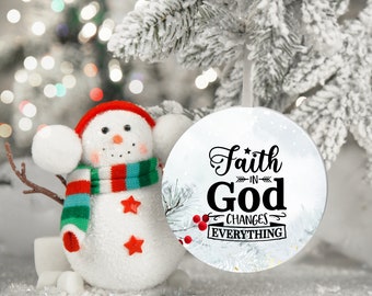 May include: A white Christmas ornament with the text "Faith in God Changes Everything" in black script. The ornament is hanging on a snowy Christmas tree branch. A snowman with a red hat and scarf is in the background.