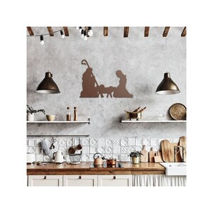 May include: A brown metal silhouette of the nativity scene with Mary, Joseph, and baby Jesus in a manger. The silhouette is mounted on a grey wall in a kitchen setting with white tiled backsplash and wooden worktops.