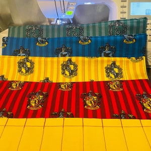 May include: Four fabric panels with the Hogwarts house crests. The panels are in the colors of each house: Gryffindor (red and gold), Hufflepuff (yellow and black), Ravenclaw (blue and bronze), and Slytherin (green and silver). The panels are stacked on top of each other, with the Gryffindor panel on the bottom and the Slytherin panel on the top.